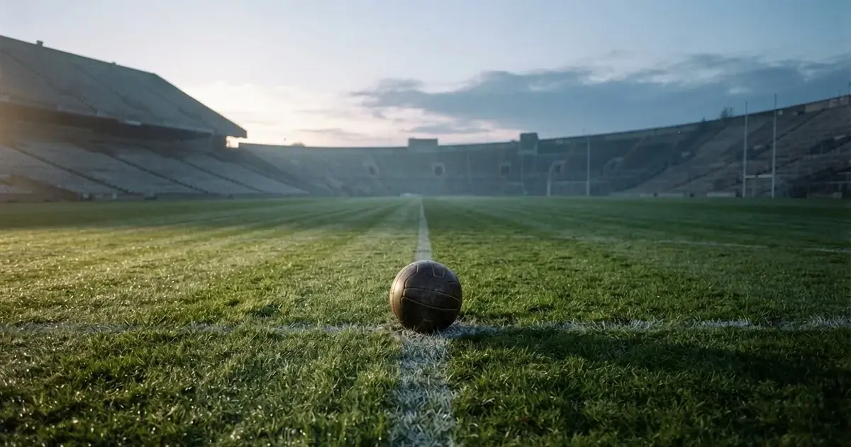 Ein Fußball liegt auf dem Anstoßpunkt eines Rasenplatzes, dahinter leere Tribünen im Licht des frühen Morgens.